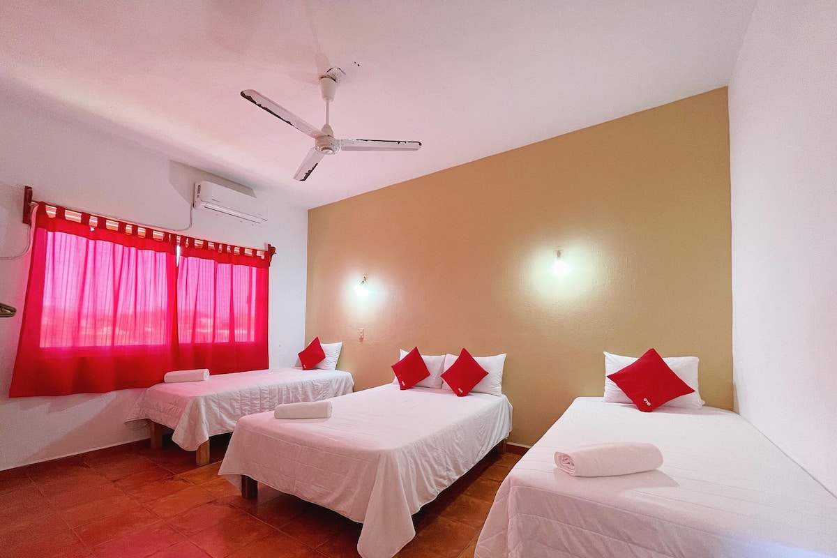 A hotel room with three single beds, each with white bedding and red pillows, a ceiling fan, and red curtains.