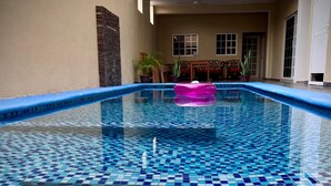 An indoor swimming pool with a blue and white tiled floor, a pink floating toy, and a view of the interior including a balcony with railings and potted plants. - Hotel y Bungalows Bugambilias (San Patricio Melaque)