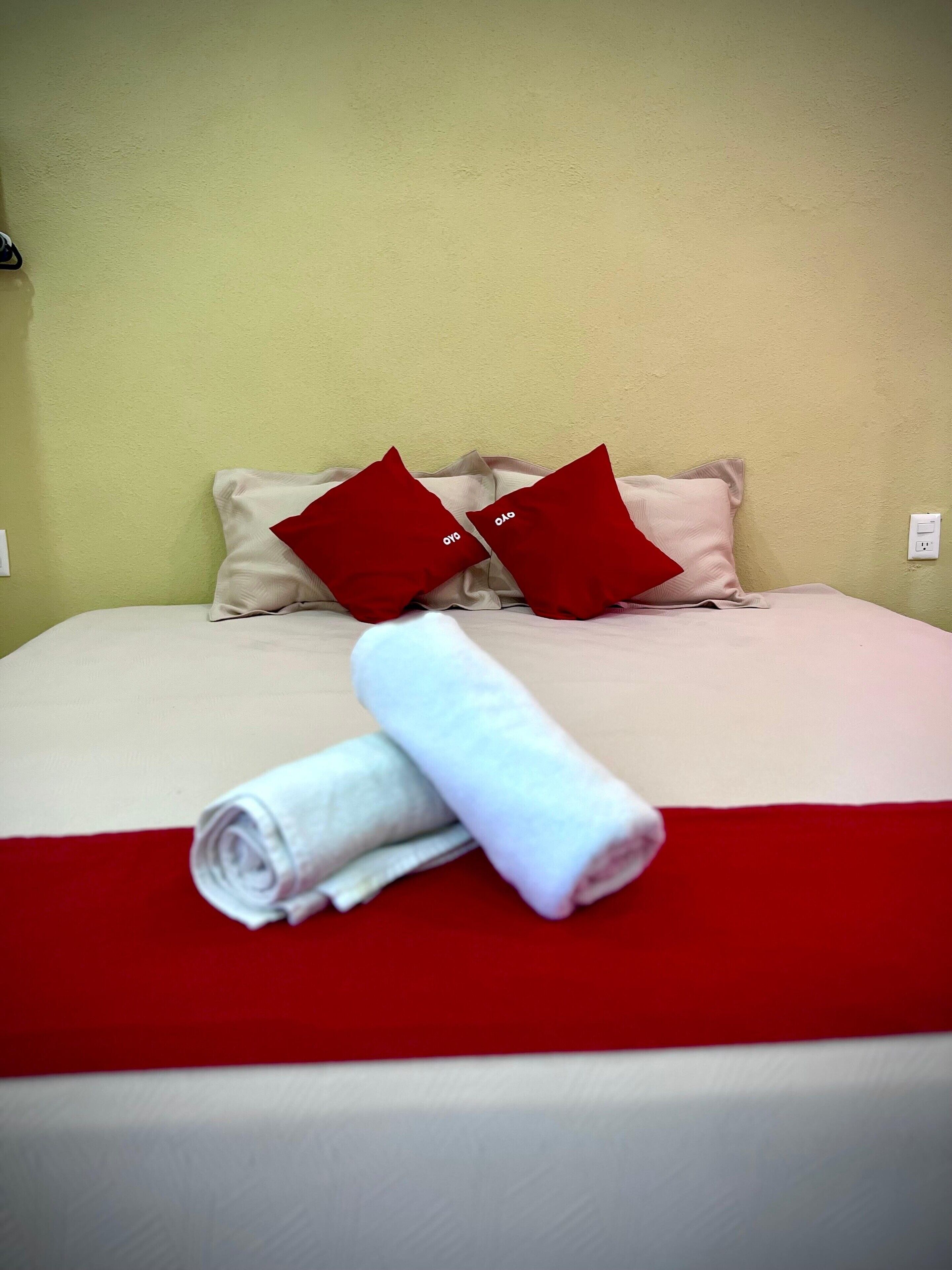 Una cama bien hecha con ropa de cama roja y beige, dos almohadas rojas y una toalla blanca enrollada sobre un corredor de cama rojo.