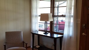 Una habitación con una silla, una mesa con una lámpara, una alfombra estampada y un ventanal con cortinas.