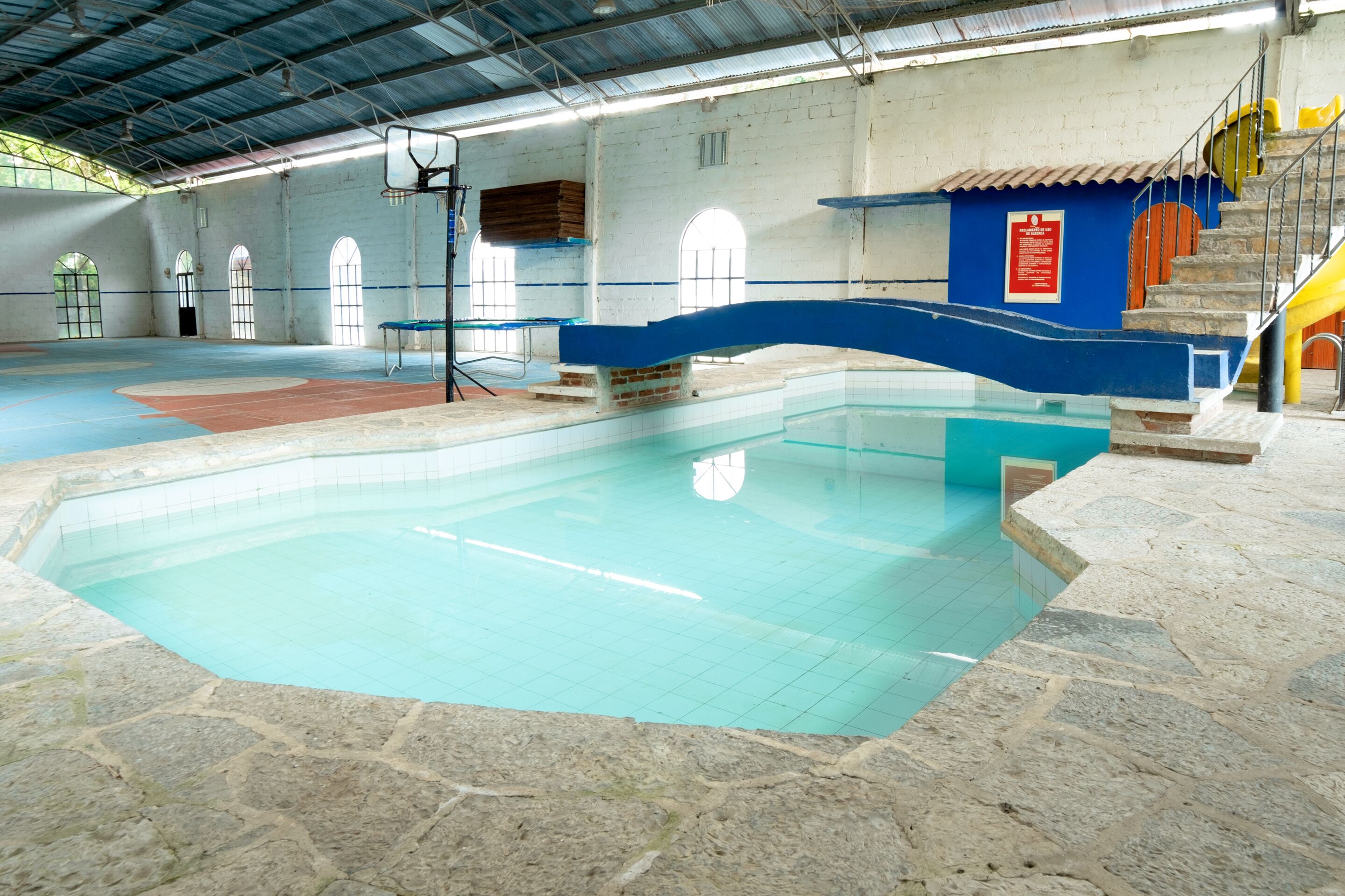 Ein hallenbad mit blauer und roter Liegefläche, Steinboden und einer Treppe, die zu einer gelben Rutsche führt.