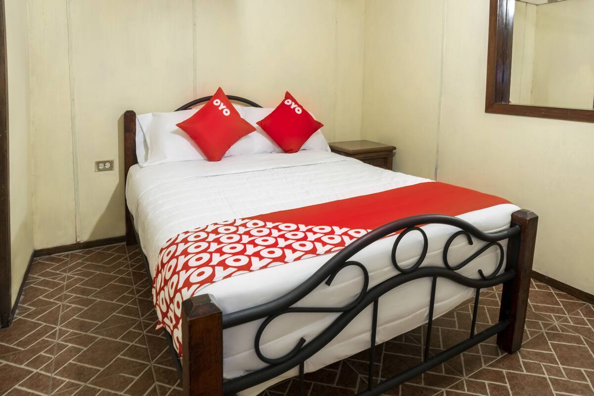 Una cama bien hecha con una colcha estampada y dos almohadas rojas, una mesita de noche de madera a su lado, y un espejo en la pared.