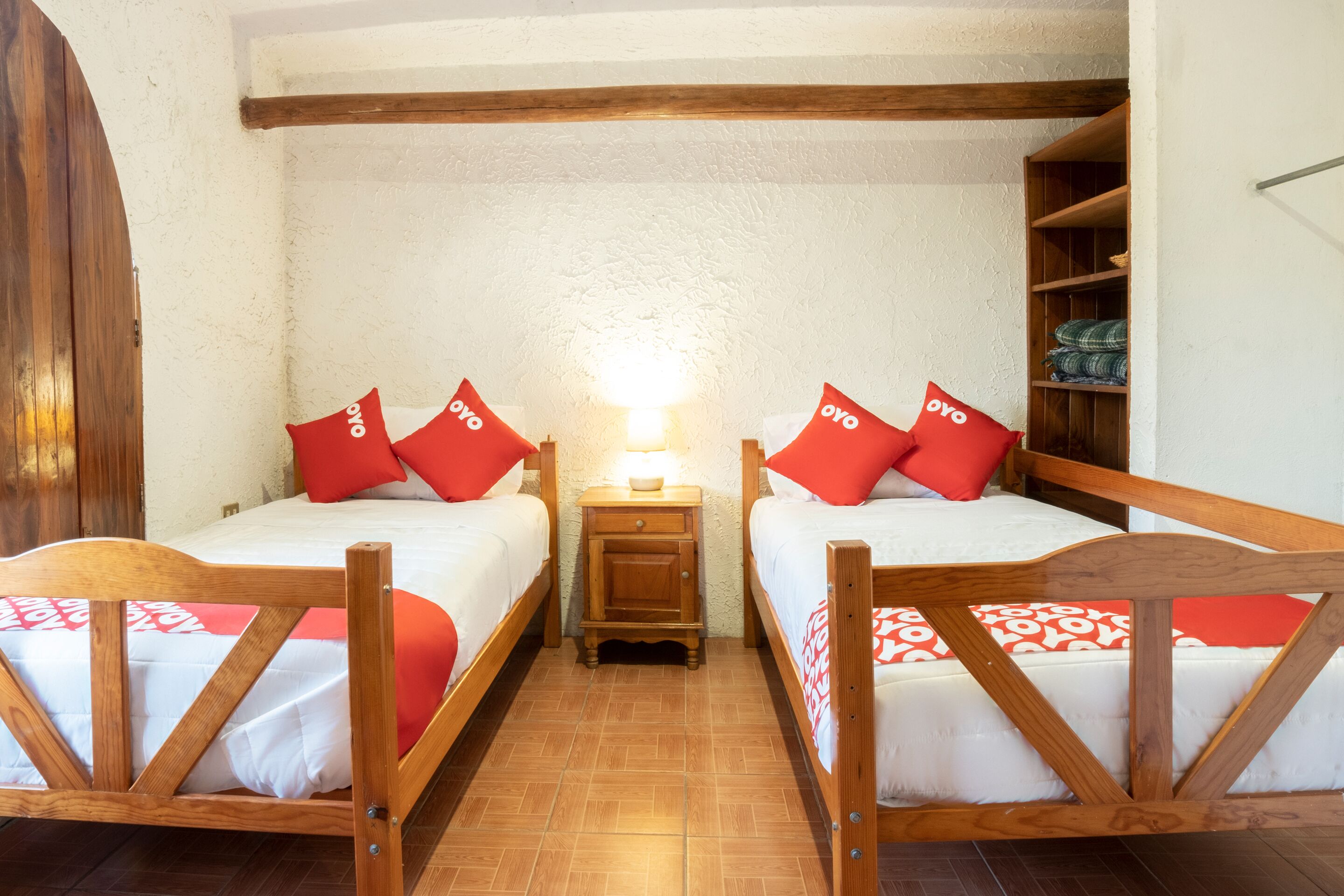 Una habitación con dos camas de madera, cada una con almohadillas rojas y una colcha blanca. Entre las camas hay una mesita de noche de madera con una lámpara. El techo es de madera y la pared es blanca.