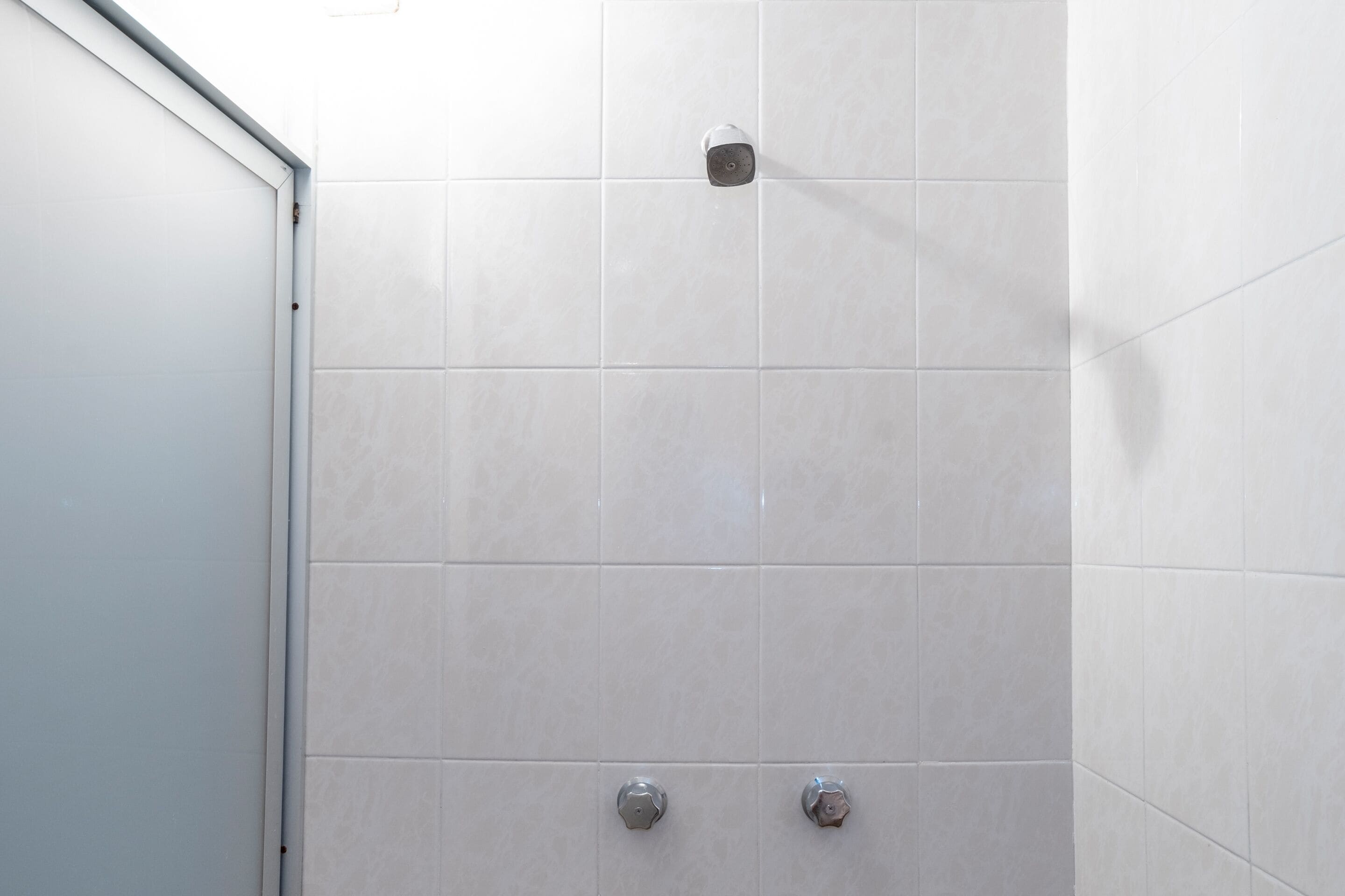 Parede de duche revestida a azulejos, com um chuveiro e duas manetes de controlo.