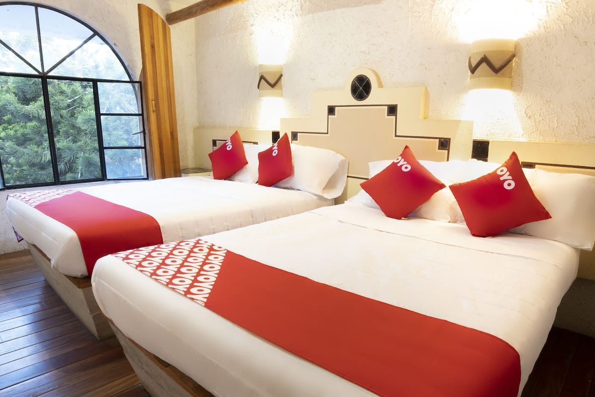 Habitación de hotel con dos camas, cada una con ropa de cama roja y blanca, un ventanal con vistas a árboles y suelo de madera.