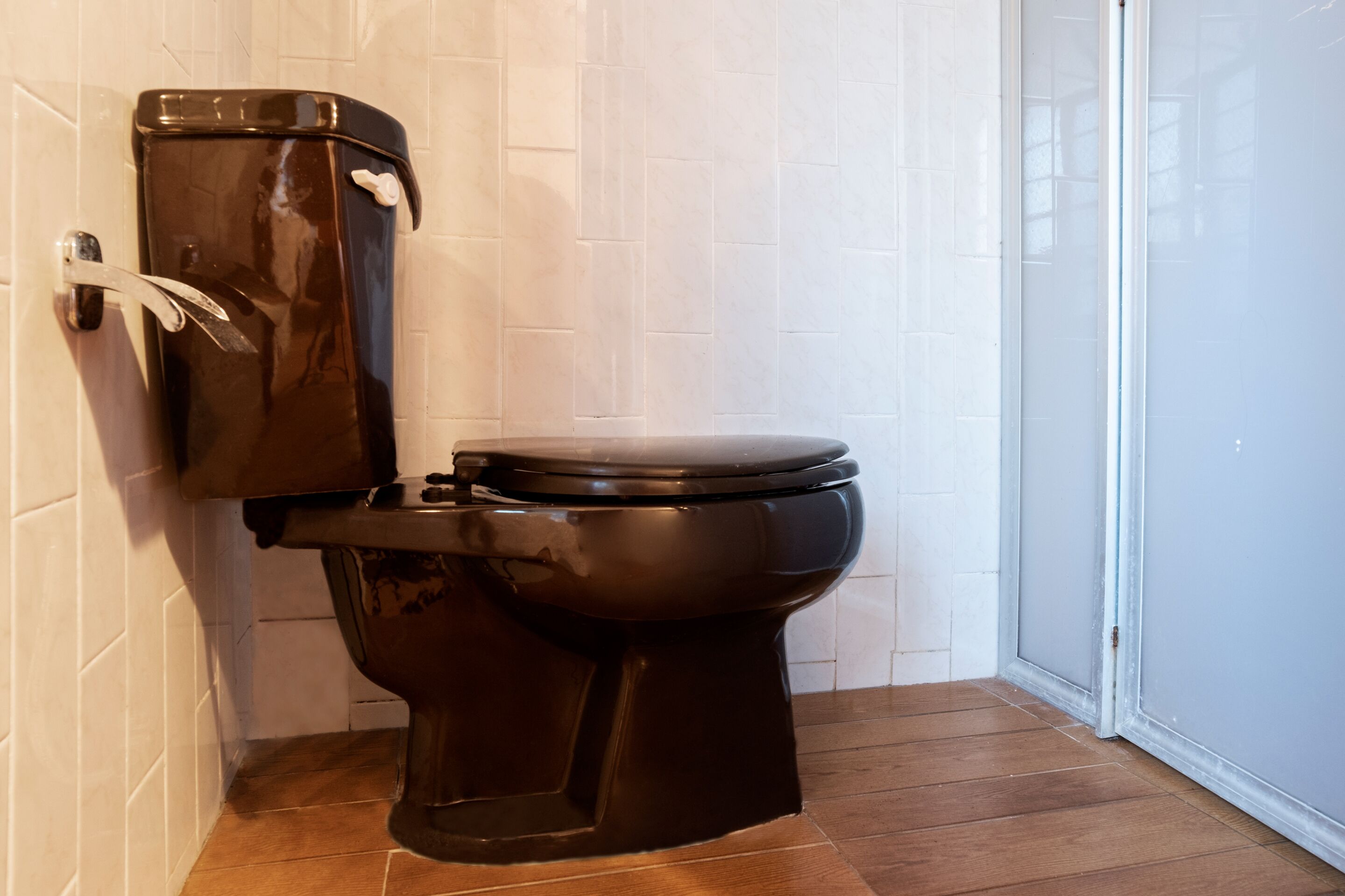 Un baño con inodoro negro, paredes de azulejos blancos y suelo de madera.