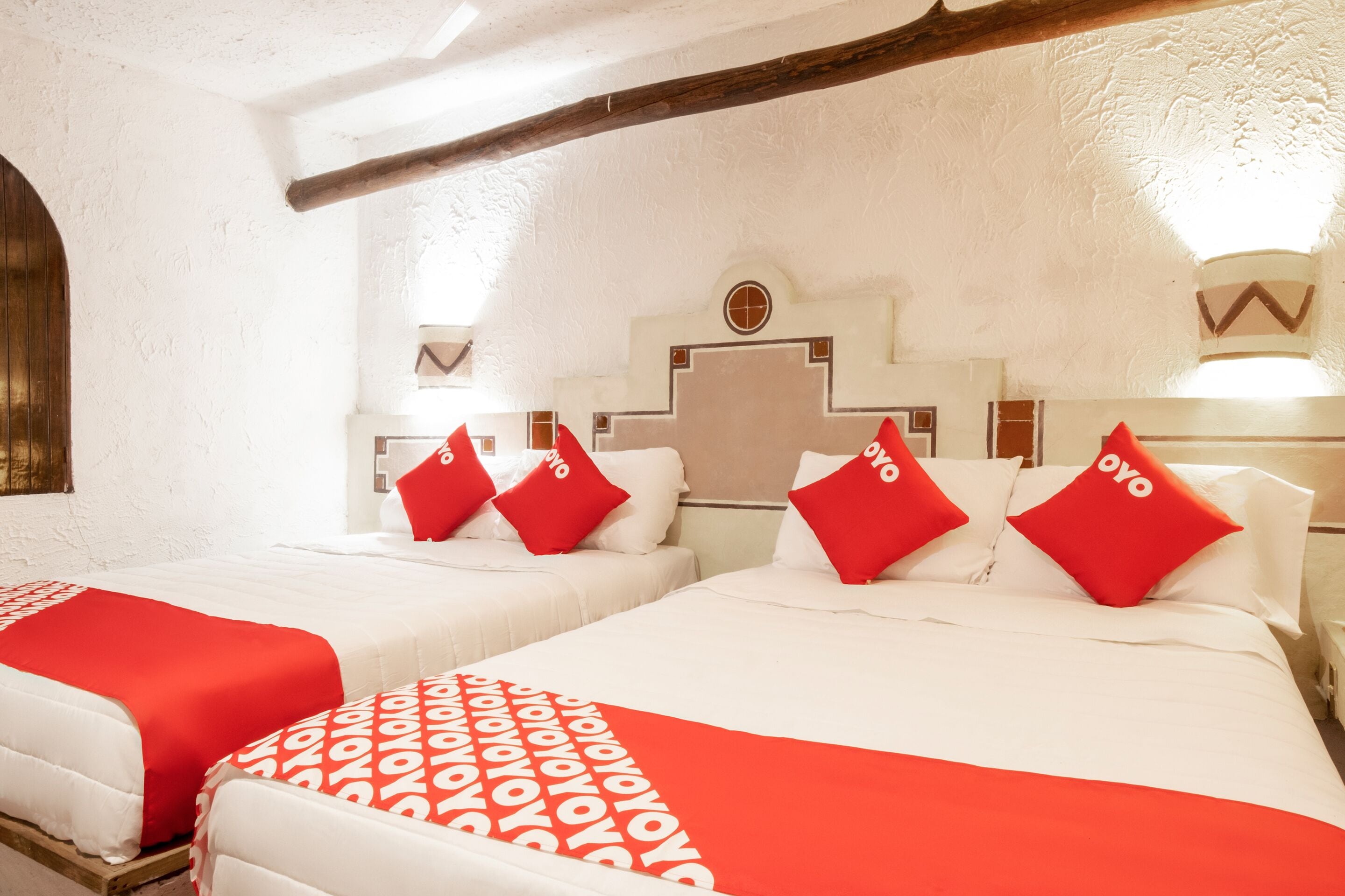A hotel room with two beds, each with red and white bedding and matching pillows. The room has a white textured wall and a wooden ceiling beam.