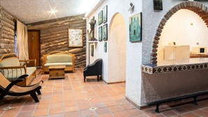 Een gezellig houten blokhut interieur met ruwe houten wanden, een betegelde vloer en een open haard. Er zijn verschillende stoelen en een bank voor zitplaatsen.