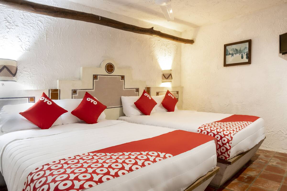 Habitación de hotel con dos camas, cada una con ropa de cama roja y blanca, cabeceros de madera y una lámpara de pared.