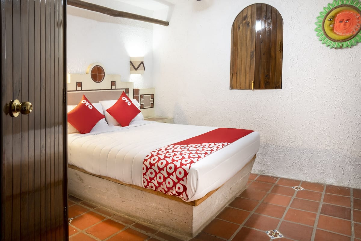 Una cama con colcha blanca y roja estampada, dos almohadas rojas y un cabecero de madera.