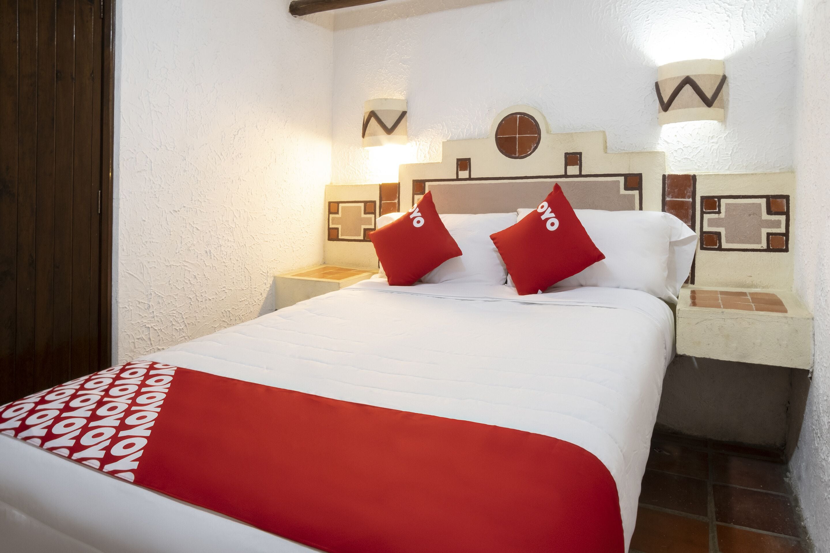 Uma cama bem arrumada com colcha vermelha e branca, dois travesseiros vermelhos e cabeceira com padrão geométrico.