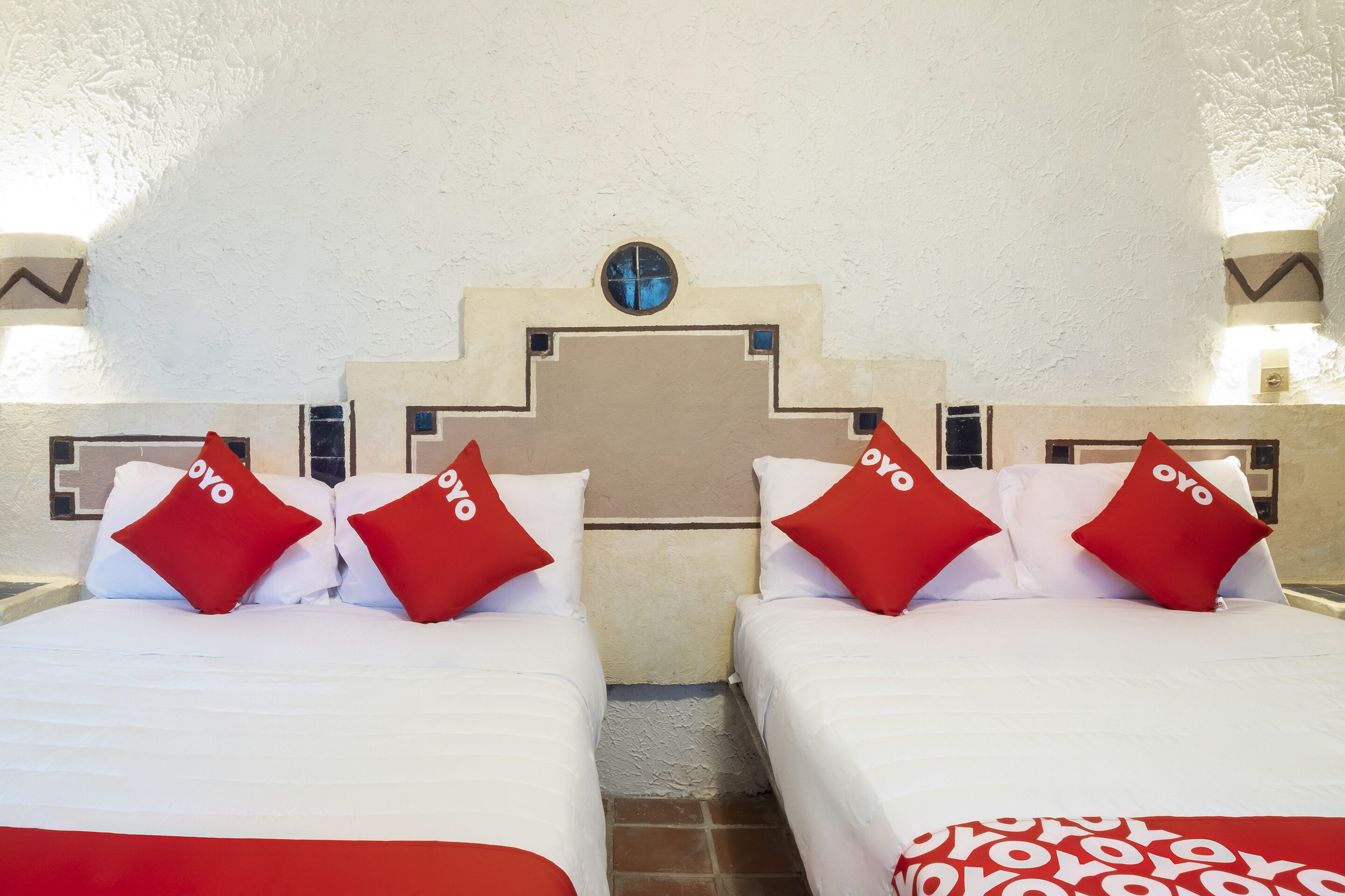 Habitación de hotel con dos camas, cada una con ropa de cama blanca y almohadas rojas. La habitación tiene una pared blanca con textura y un techo decorativo con un ventanal circular.
