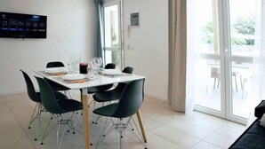 Uma sala de jantar com uma mesa posta para quatro pessoas, cadeiras pretas, uma TV de tela plana na parede e uma porta de vidro deslizante que dá para uma área externa.