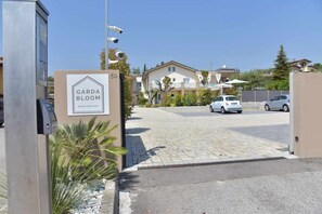 A gated entrance to a residential area with a sign for 'Garda Bloom Holiday Apartments' and a parking lot with cars.
