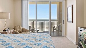 A hotel room with a bed, a desk with a lamp, a chair, a dresser, and a balcony with a view of the beach. - Oceanfront Penthouse Villa w/ Balcony, Hilton Head (Hilton Head Island)