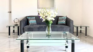 Una sala de estar moderna con una mesa de centro de vidrio, sofá gris, cojines estampados y un jarrón con flores.