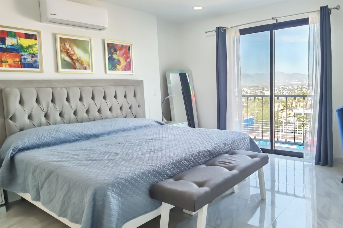 A bedroom with a large bed, a bench, a bedside table, a lamp, and a view of a balcony with a railing.