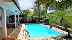 Ein Pool mit einer terrasse aus Steinplatten, umgeben von tropischen Pflanzen und einem überdachten Außenbereich mit blauen Akzenten.