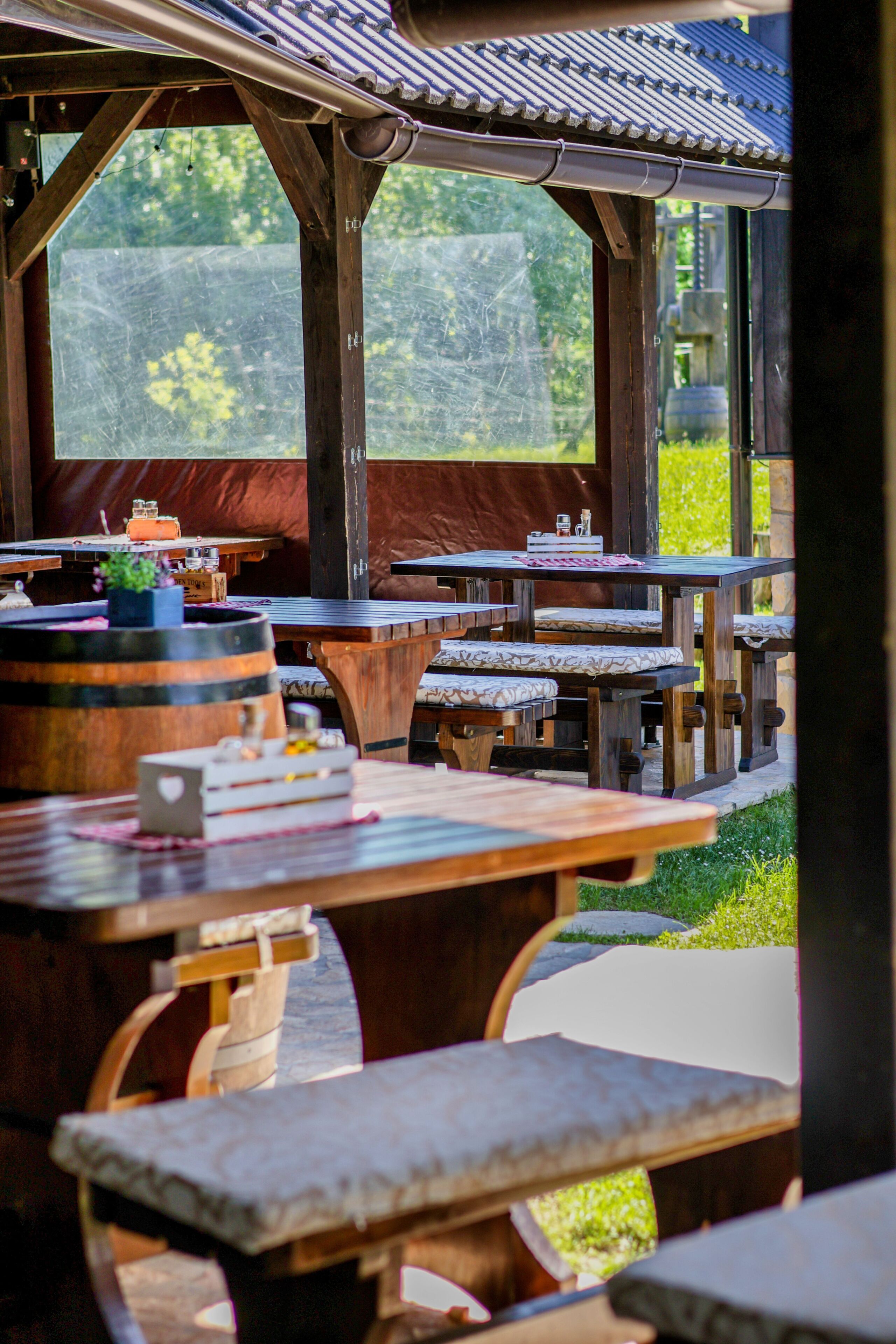 An outdoor dining area with wooden tables and benches, a covered roof, and a view of greenery.