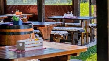 An outdoor dining area with wooden tables and benches, a covered roof, and a view of greenery.