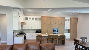 A modern kitchen with white cabinets, a central island with bar stools, and a brown leather sofa in the living area.