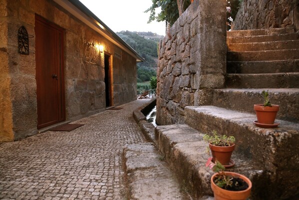 A stone building with a wooden door and a lit wall sign, a cobblestone path, and a stone staircase with potted plants.