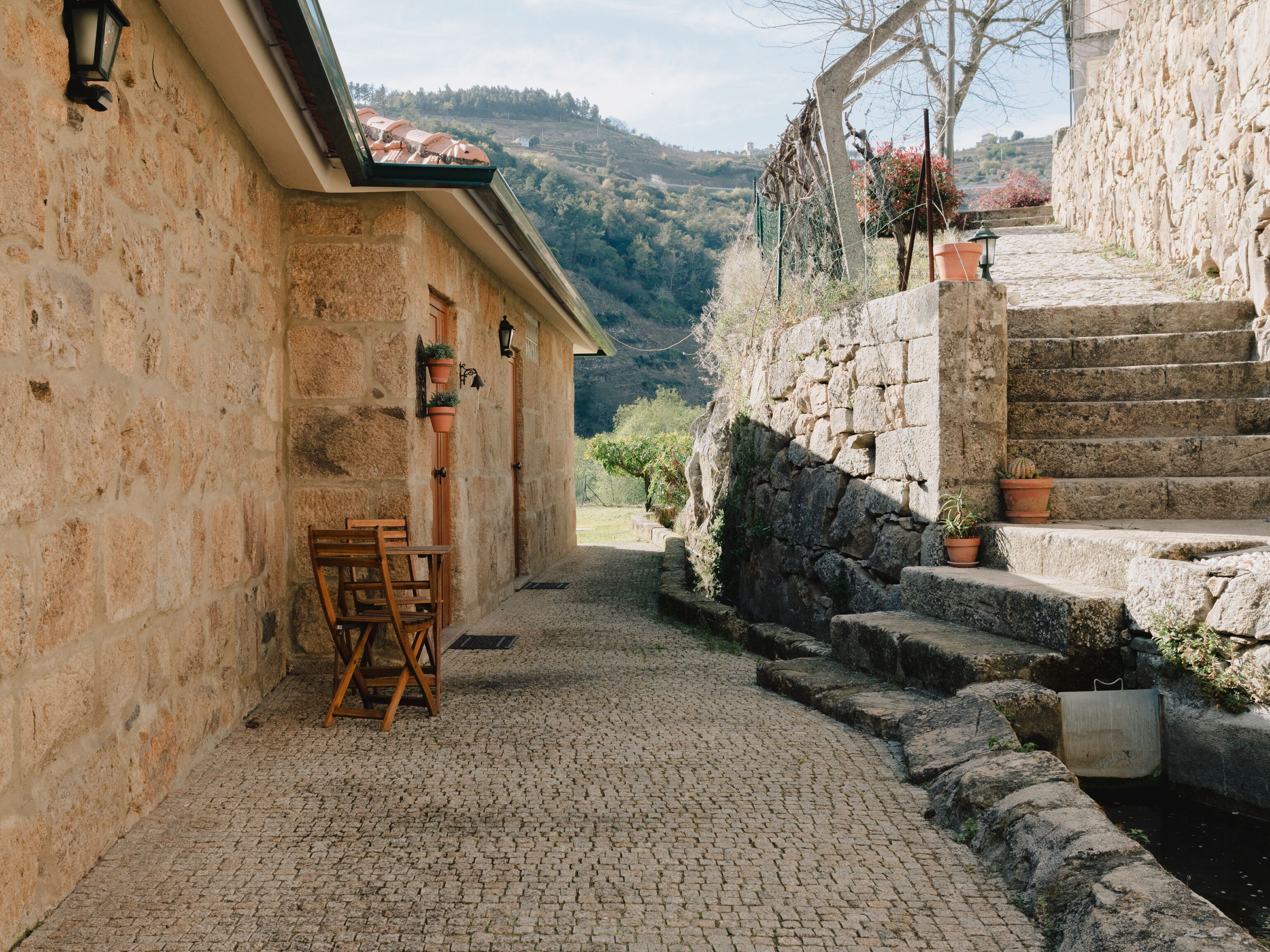 A stone building with a cobblestone courtyard, wooden chairs, and a view of a hillside.