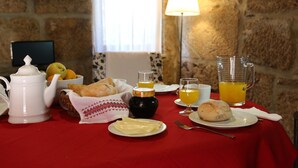 Una mesa de desayuno con mantel rojo, tetera blanca, un cuenco de fruta, cesta de pan, una fuente de tortitas y vasos de zumo de naranja.