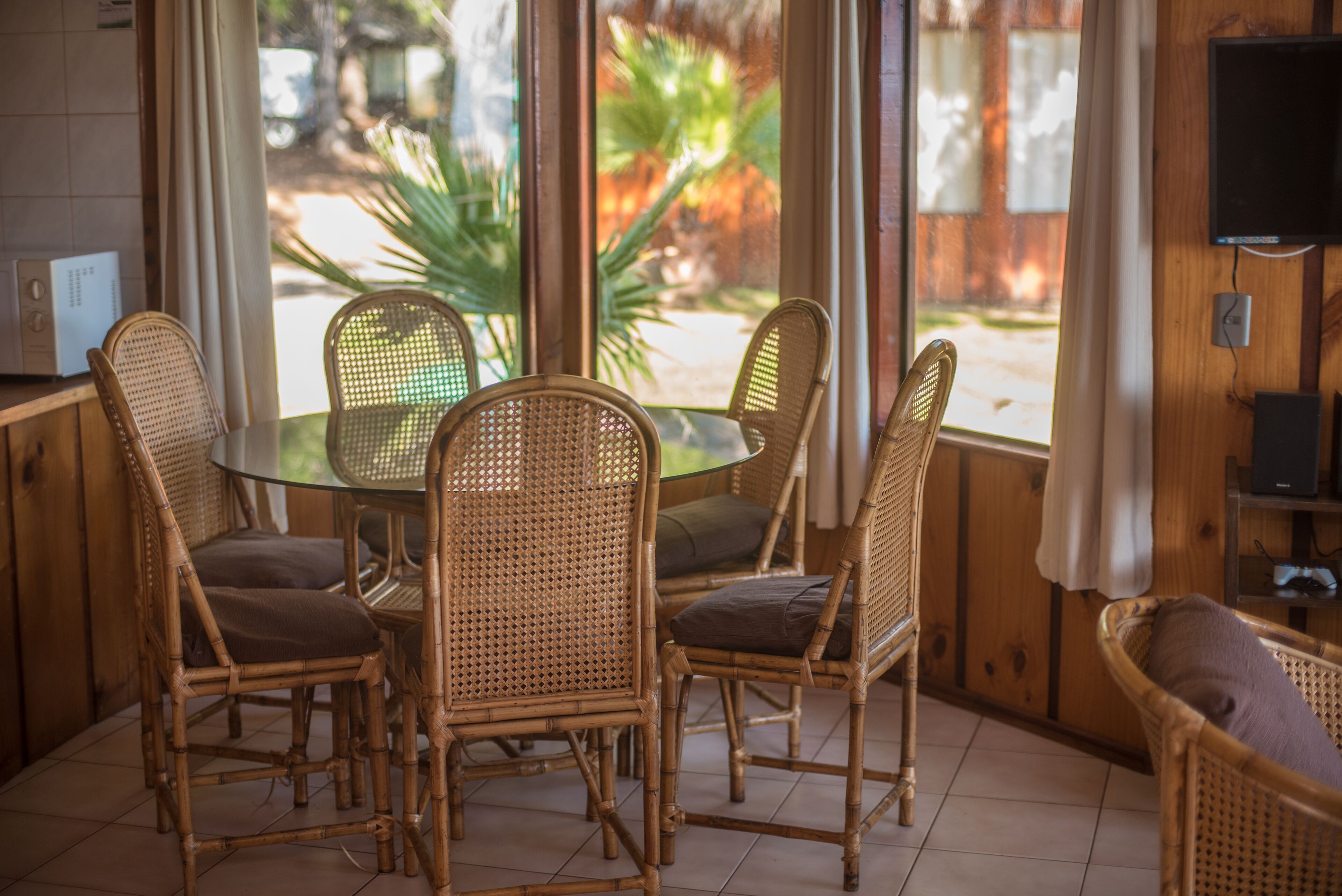 A dining area with wicker chairs and a glass table, large windows with curtains, and a television on the wall.