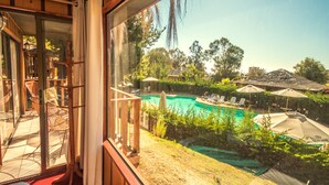 Vue depuis une chambre dotée d’une grande fenêtre donnant sur une piscine, du mobilier d’extérieur et la végétation environnante.