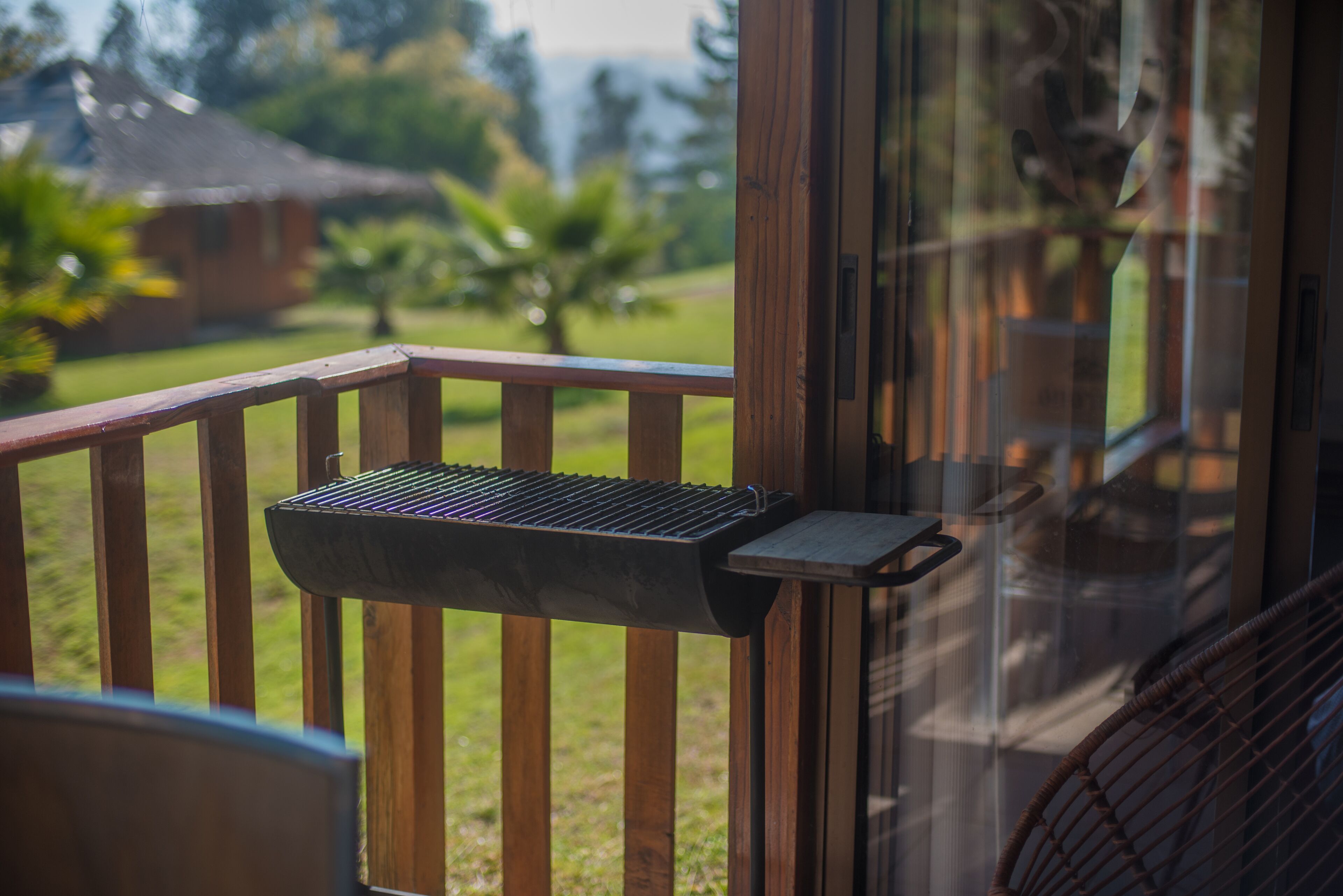 A wooden deck with a grill attached to the railing, overlooking a lush garden and a house in the background.