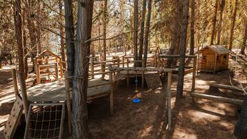 A forested playground with wooden climbing structures, a small wooden house, and a rope swing.