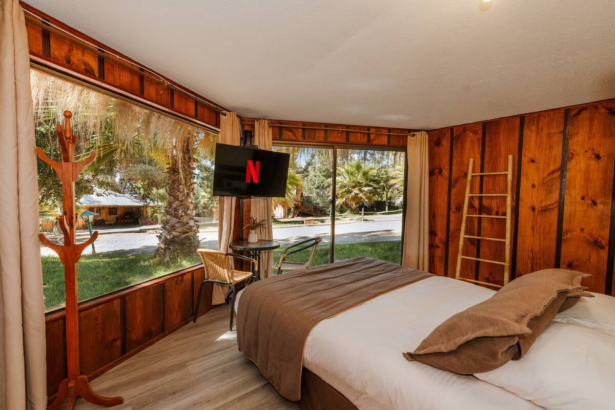 Un dormitorio con una cama grande, un televisor montado en la pared, una silla de madera y vistas al exterior.