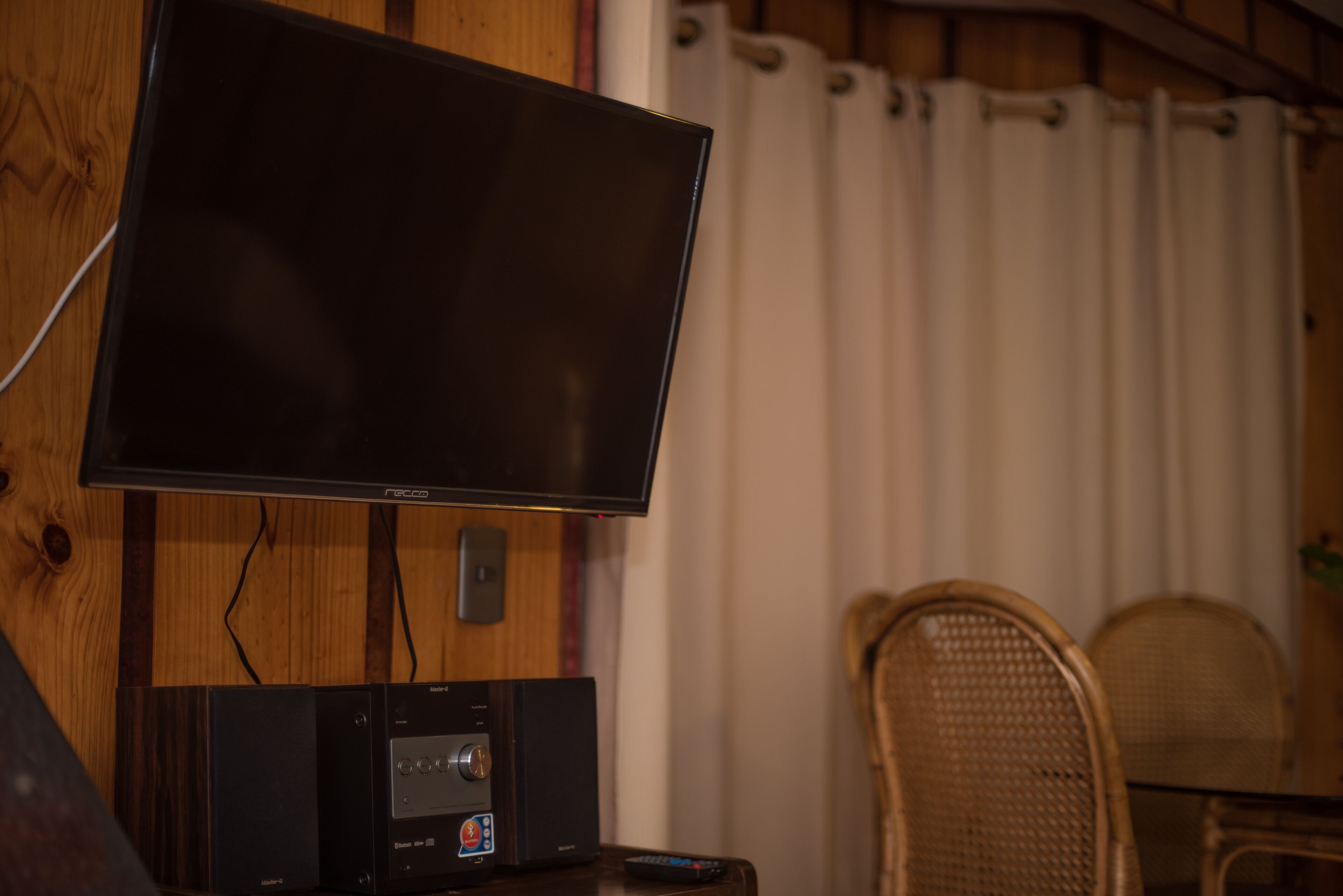 A flat-screen TV mounted on a wooden wall, a sound system below it, and wicker chairs in the background.