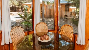 Un intérieur en bois avec une table en verre entourée de chaises en osier, un petit objet décoratif posé sur la table, et une vue sur un espace extérieur agrémenté de plantes et d’une clôture en bois.