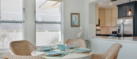 Un comedor con sillas de mimbre, una mesa redonda dispuesta con platos y vasos, y vista a la cocina con refrigerador y armarios.