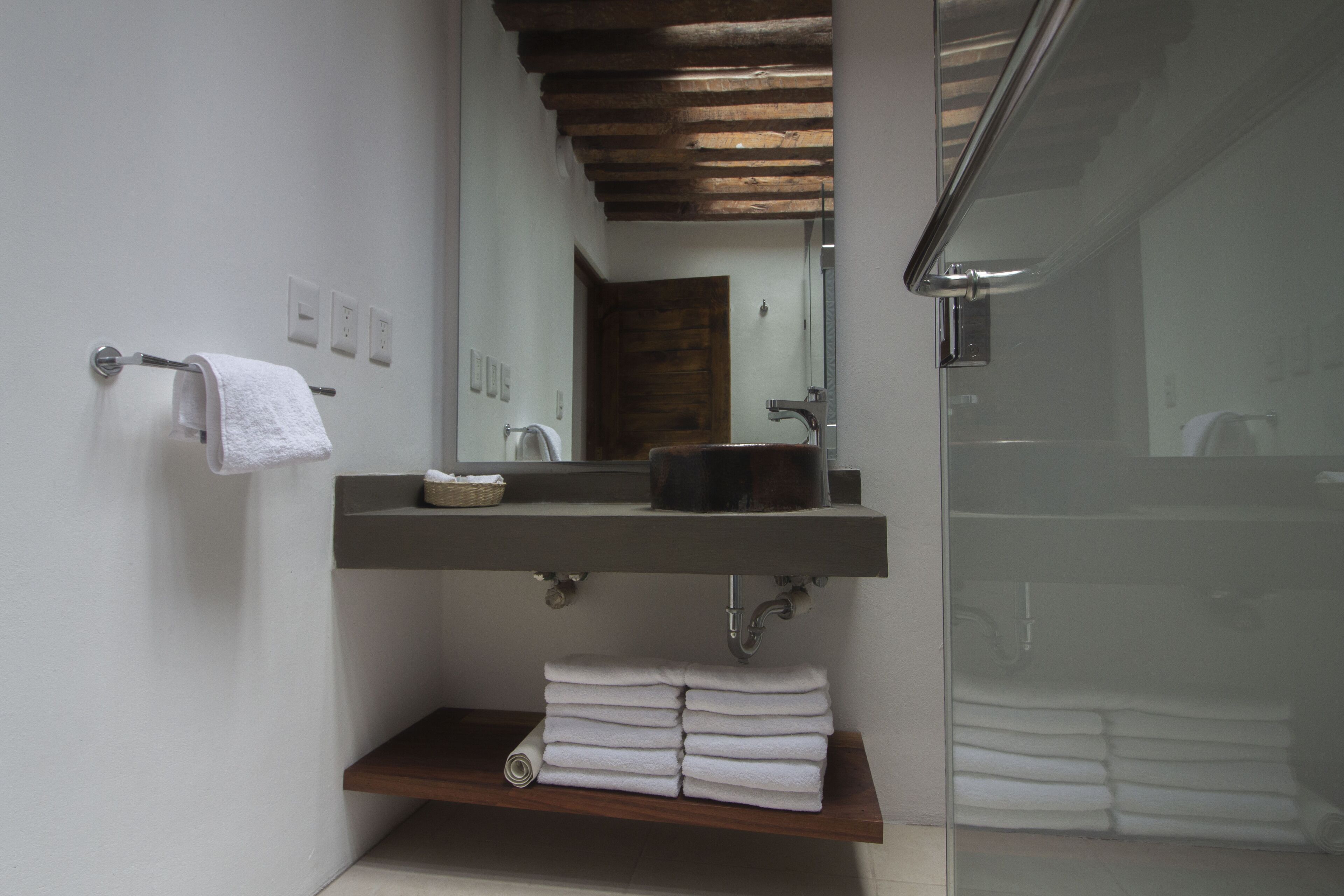 A modern bathroom with a wooden shelf holding towels, a sink with a mirror above, and a glass shower enclosure.