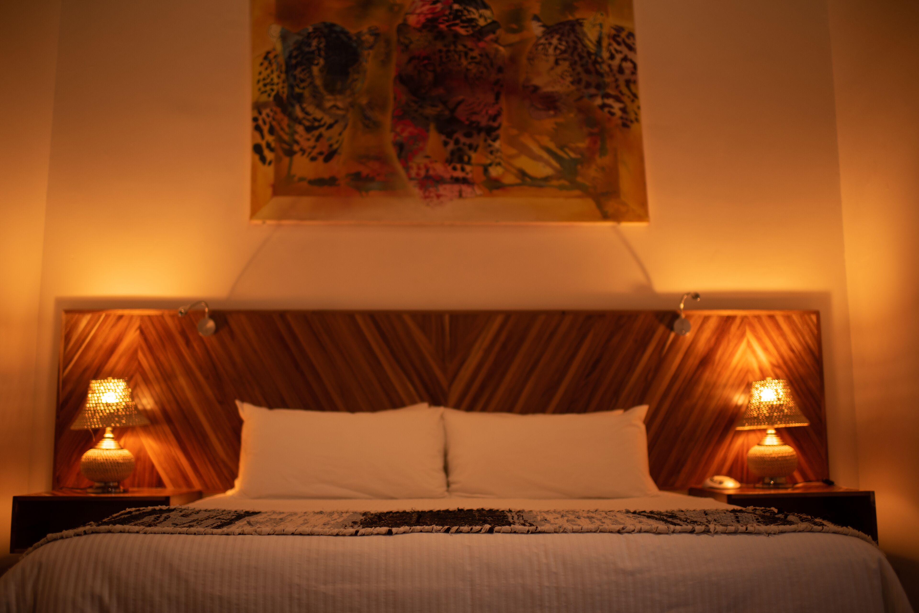 A hotel room with a wooden headboard, two bedside lamps, a bed with white linens, and a painting on the wall.