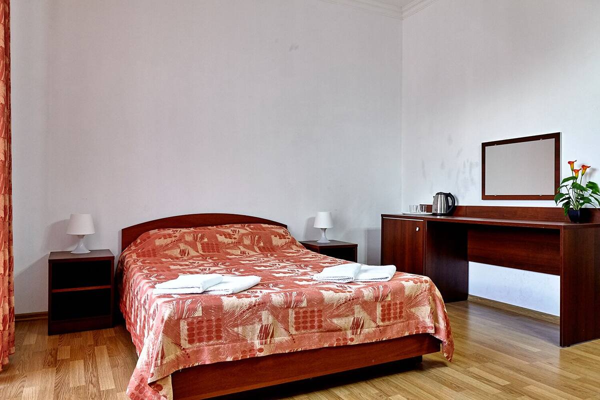 Una habitación de hotel con una cama de madera, dos mesitas de noche con lámparas, un escritorio con espejo y una planta en maceta.