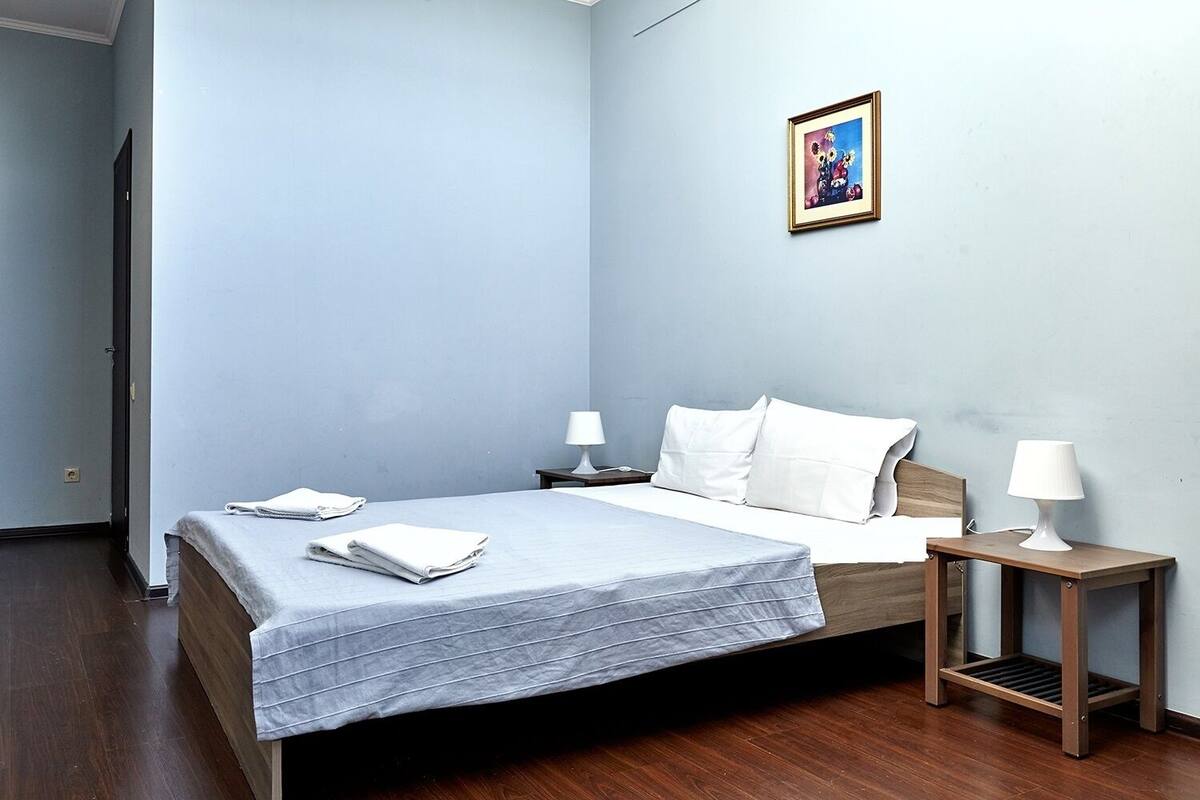 Una cama bien hecha con sábanas blancas y una manta gris, dos mesitas de noche con lámparas y un cuadro en la pared.