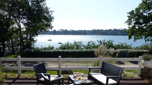 Un espace extérieur aménagé avec deux chaises et une table où sont disposés des plats, donnant sur un lac où l’on peut voir des bateaux.