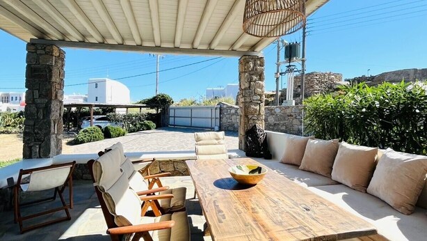 An outdoor patio with a wooden table, cushioned seating, a hanging birdcage, and stone pillars. - Drafies House (Mykonos)