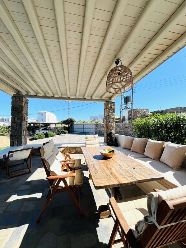 An outdoor patio with a wooden table, cushioned seating, a hanging birdcage, and stone pillars.
