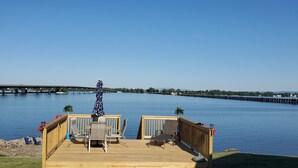 A wooden deck with outdoor furniture overlooking a calm body of water, a bridge in the background, and a pier extending into the water. - Year-Round: Amazing Sunrises, 150ft Shore, Private Boat Launch/Dock, Hot Fishing (Alburg)