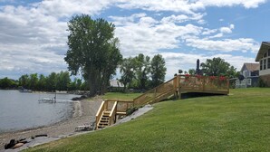 A wooden deck leading to a house by a lake with a dock and a large tree. - Year-Round: Amazing Sunrises, 150ft Shore, Private Boat Launch/Dock, Hot Fishing (Alburg)