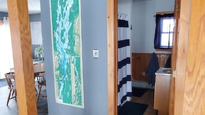 A wooden staircase leading to a room with a map on the wall, a bathroom with a shower curtain, and a kitchen area visible through an open door. - Year-Round: Amazing Sunrises, 150ft Shore, Private Boat Launch/Dock, Hot Fishing (Alburg)