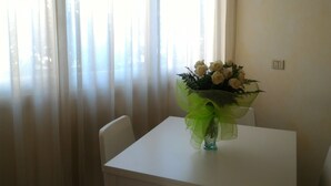 A dining table with a vase of white flowers, a clock on the wall, and sheer curtains.