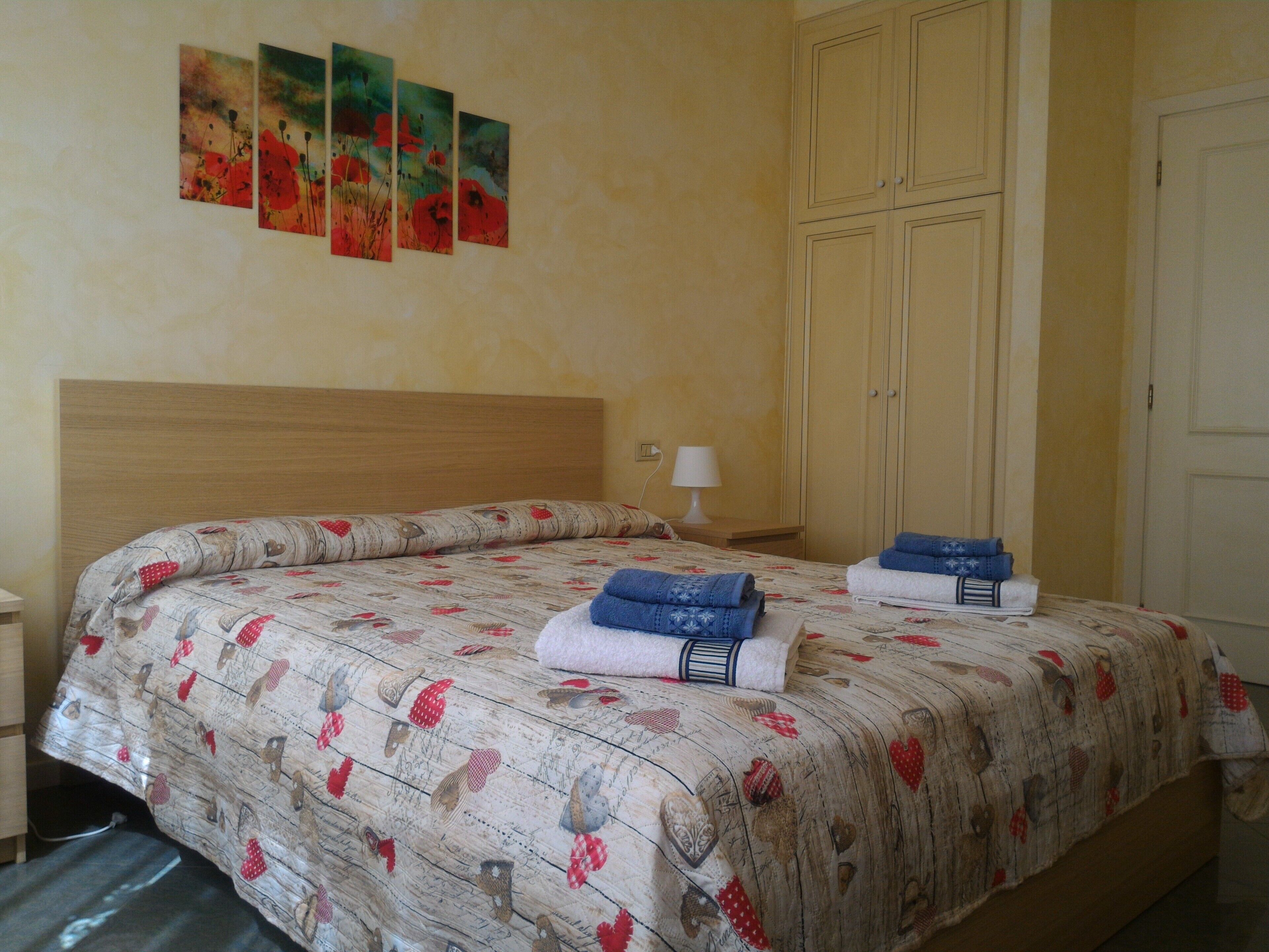 Un dormitorio con cabecera de madera, cama con colcha de motivos florales, dos toallas dobladas, una lámpara de noche y un armario.