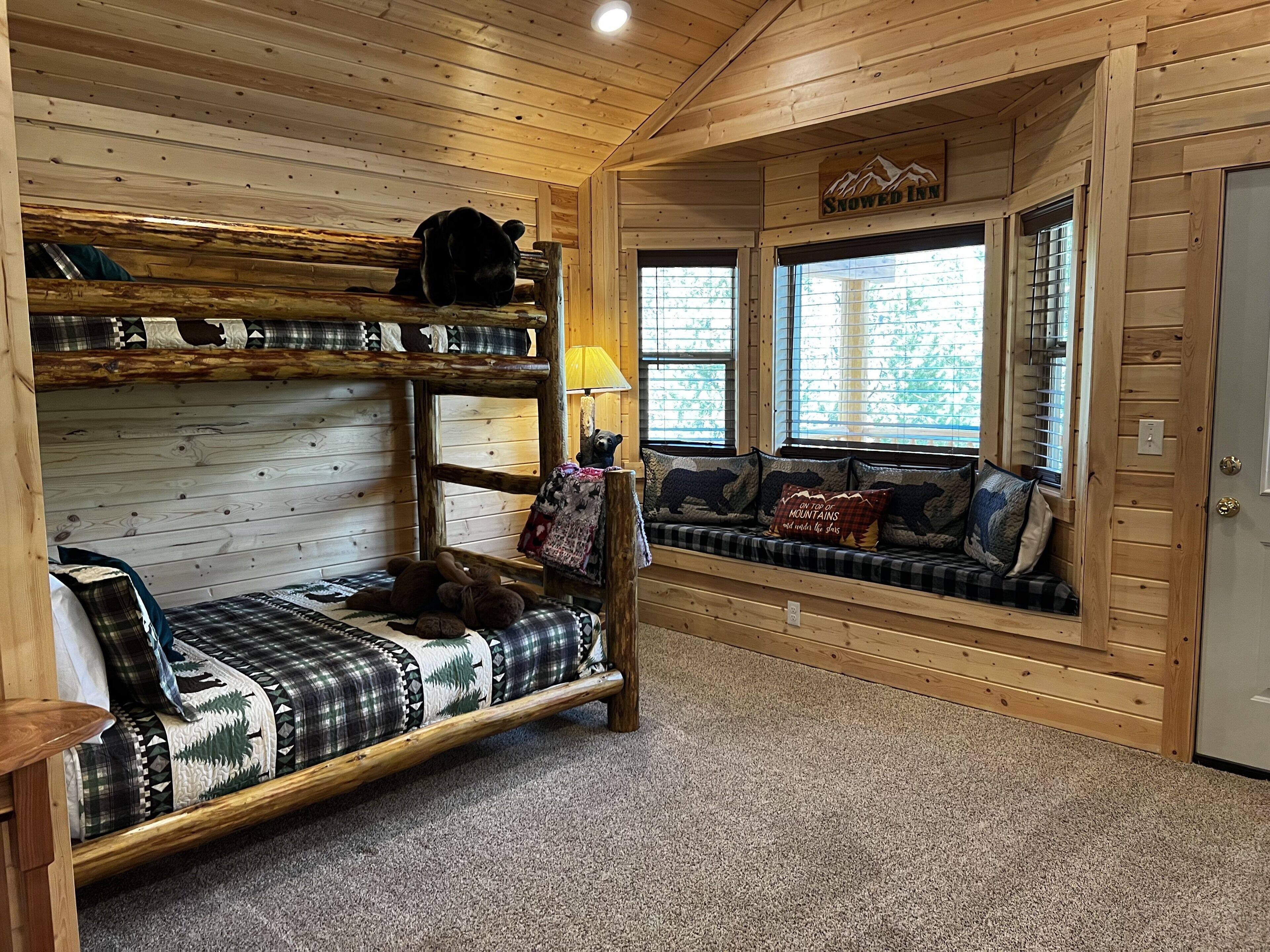 Un intérieur de chalet chaleureux avec un lit superposé, un canapé agrémenté de coussins à motifs de montagne et une fenêtre avec des stores.
