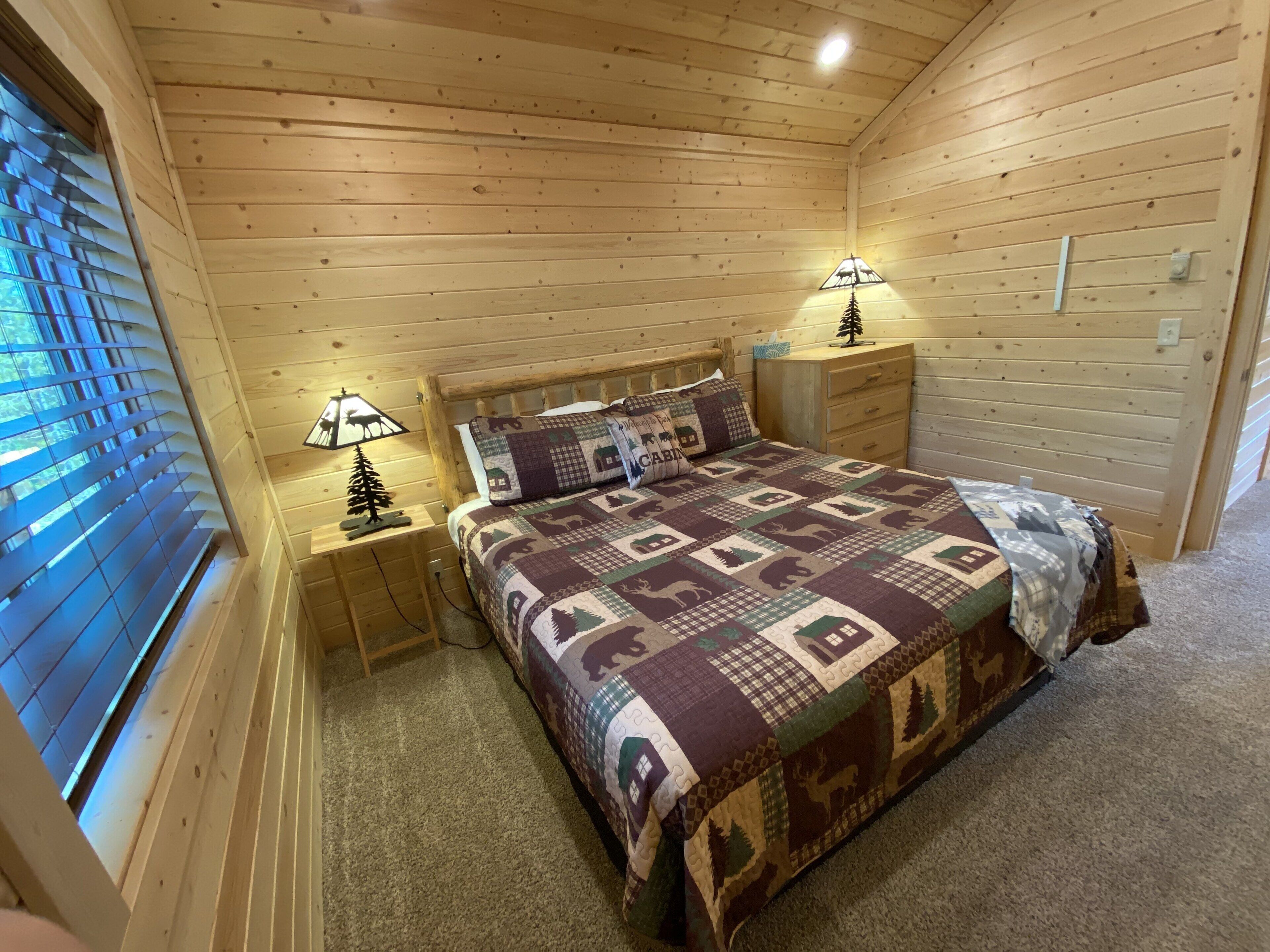 A cozy cabin-style bedroom with a wooden ceiling, a quilted bedspread, two bedside tables with lamps, and a window with blinds.